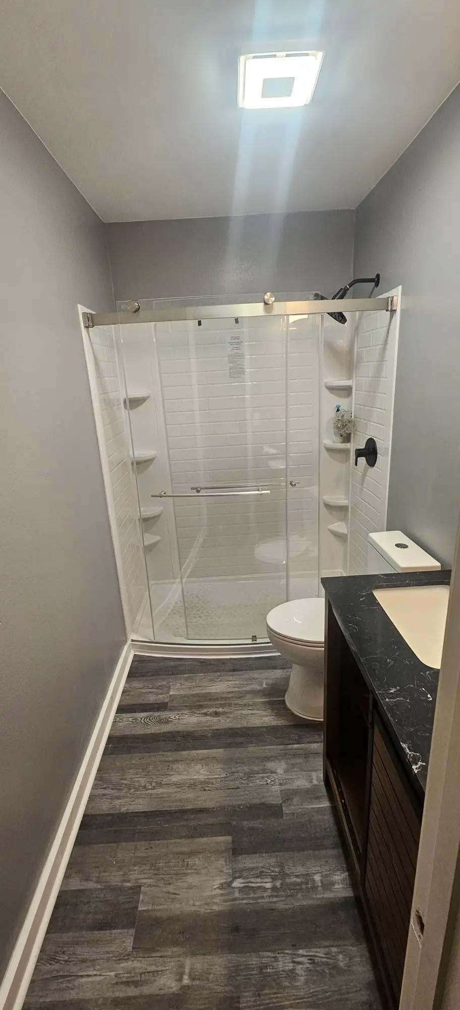 After: neo-angle shower with a sliding glass door and a stone-topped vanity on gray plank flooring.