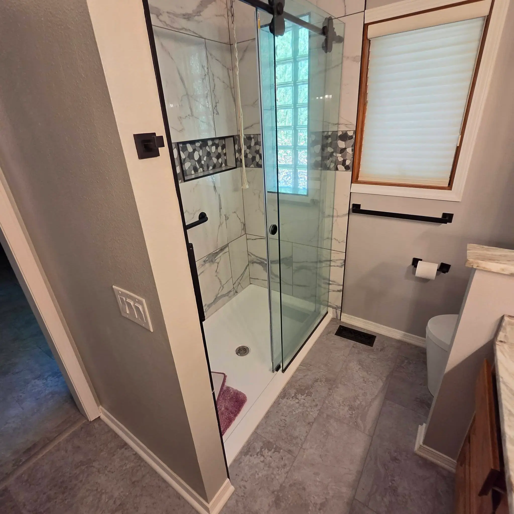 After: rebuilt walk-in shower tiled in marble-look porcelain with a pebble mosaic band and a frameless glass door.