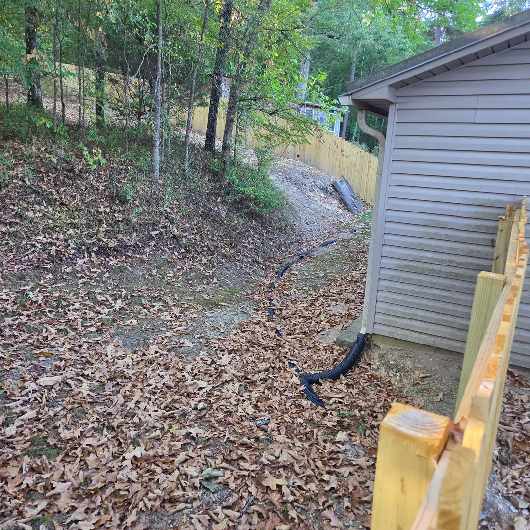 Before: a bare wash beside the house where roof runoff cut a channel, with the downspout extension lying loose on the leaves.