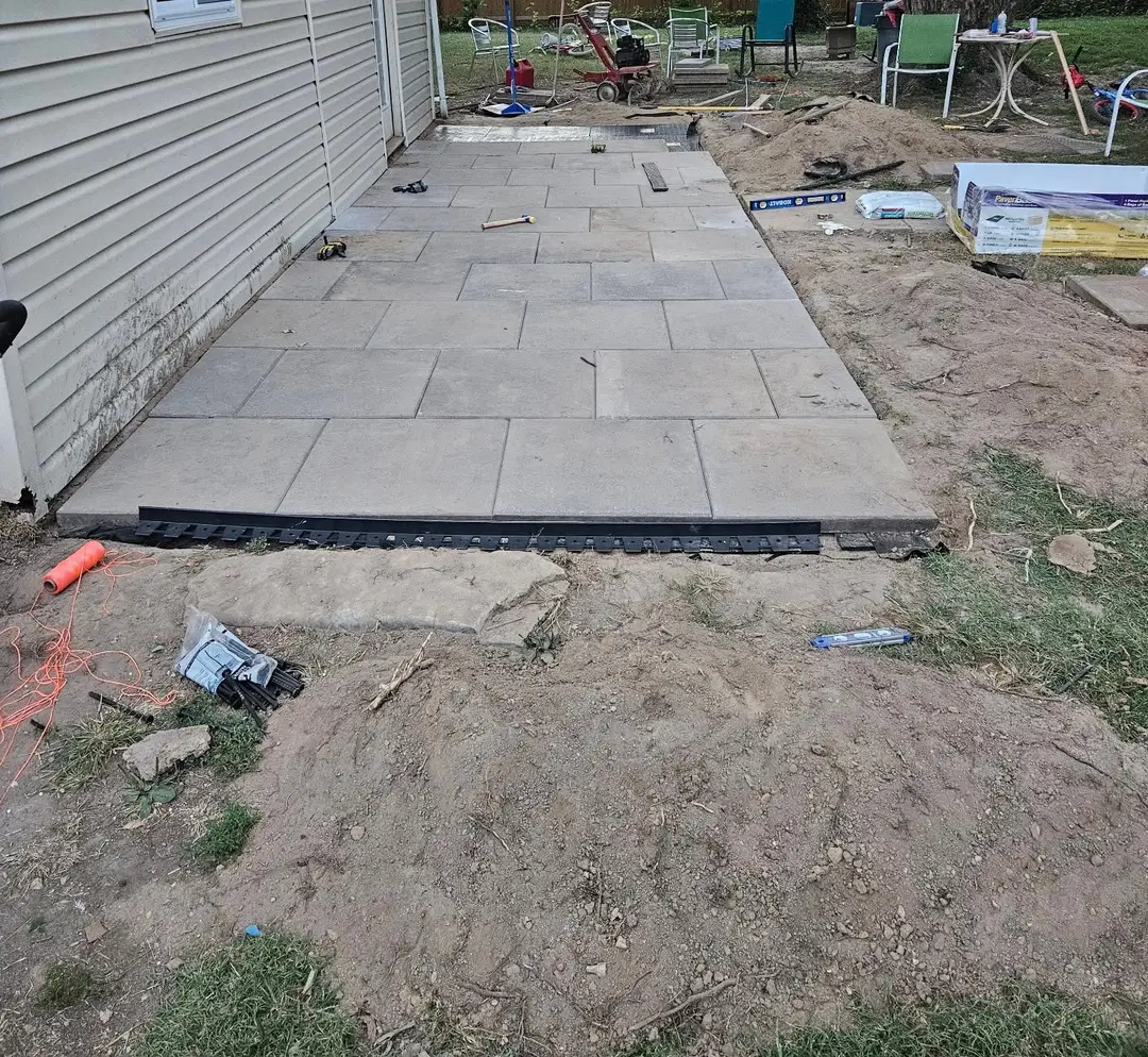 Before: half-laid paver patio along an outbuilding wall, with the edge restraint set and bare dirt beyond it.