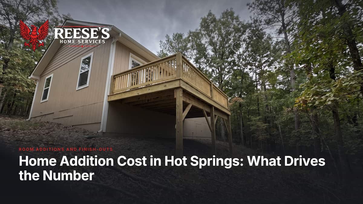 Tan-sided addition on a wooded lot, with a raised wood deck and railing standing on tall posts.